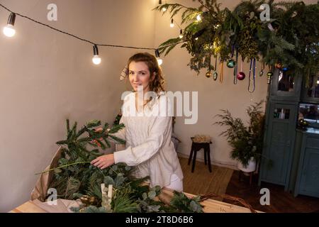 Jeune femme en pull tricoté léger choisit des aiguilles de pin naturel vert pour la couronne de Noël faite à la main sur la table en bois. Une fille millénaire fait de la décoratio Banque D'Images