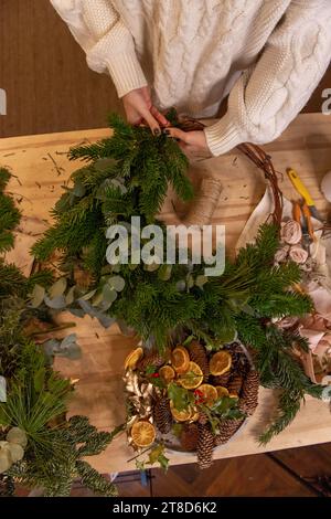 Vue de dessus composition de mains femelles faisant couronne de Noël à partir de branches naturelles d'épinette, de pin, d'eucalyptus. Fille enroule des brindilles vertes sur la base Kraft Banque D'Images