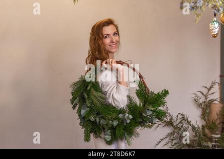 Portrait de jeune femme tenant une couronne de Noël verte asymétrique faite de matériaux naturels sur l'épaule dans les mains. Fille en beige tricoté avec Homemad Banque D'Images