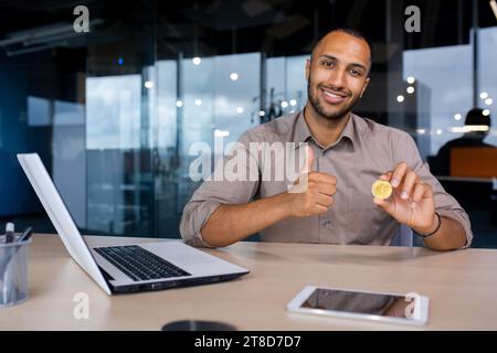 Heureux homme d'affaires afro-américain assis dans le bureau tenant le bitcoin virtuel de crypto-monnaie montrant comme pouce vers le haut. Banque D'Images