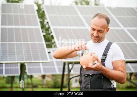 Homme mettant de l'argent dans la tirelire sur fond de panneaux solaires. Homme adulte faisant des investissements dans l'avenir. Homme compétent investissant dans des sources d'énergie alternatives. Banque D'Images