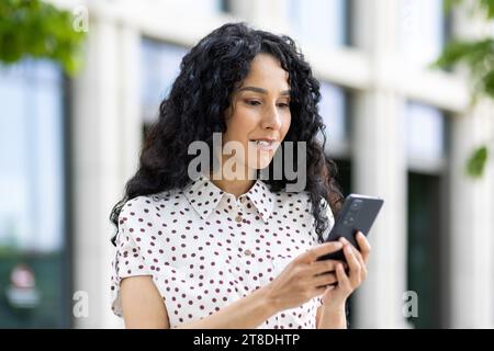 Une jeune belle femme se promène dans la ville avec un téléphone dans les mains, sourit contentement, utilise une application pour smartphone, navigue sur des pages Internet, tape un message texte, les réseaux sociaux. Banque D'Images