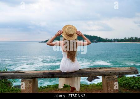 Jeune femme en robe blanche est assise sur un banc rustique au bord de la mer, contemplant les vagues. Vue arrière de la femme dans le chapeau de paille bénéficie du paysage de l'océan, la solitude Banque D'Images