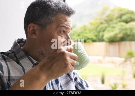 Homme biracial senior réfléchi buvant du café à la fenêtre à la maison Banque D'Images