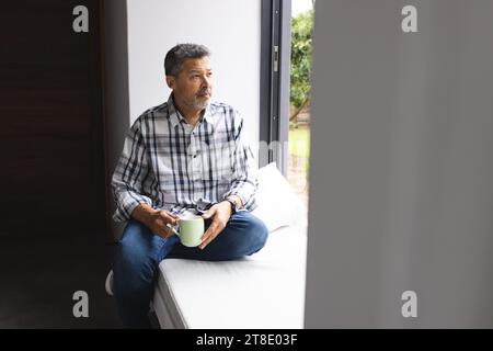 Homme biracial senior réfléchi buvant du café à la fenêtre à la maison Banque D'Images