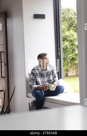 Homme biracial senior réfléchi buvant du café à la fenêtre à la maison Banque D'Images