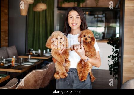 Belle femme avec deux chiens dans le restaurant. Portrait de modèle féminin souriant, regardant la caméra et tenant les animaux sur les mains à l'intérieur. Propriétaire avec caniche p Banque D'Images