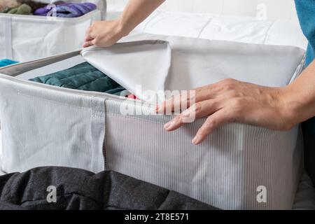 Photo horizontale, main de femme fermant la fermeture éclair d'une boîte en tissu, avec des manteaux à l'intérieur, sur un lit, collectant des vêtements d'hiver au changement de saison. Banque D'Images