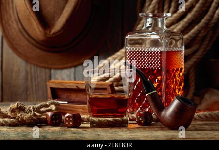 Whisky, pipe à fumer et dés sur une vieille table en bois. En arrière-plan se trouve une corde de chanvre et un chapeau d'homme. Banque D'Images