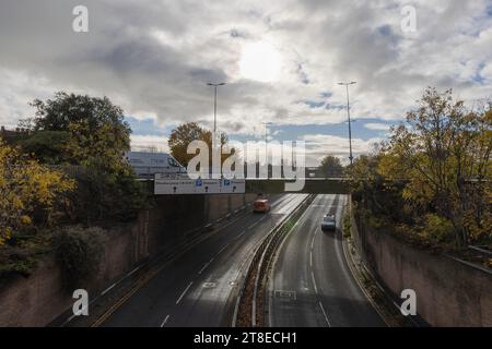 Chaussée mouillée Queensway à Southend-on-Sea, les voitures passent sous le pont avec des panneaux de stationnement. Les arbres d'automne et les nuages ensoleillés encadrent la scène urbaine. Banque D'Images