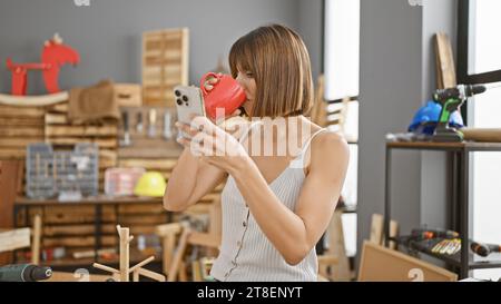 Époustouflante jeune femme hispanique charpentière tapant intensément sur un smartphone tout en savourant son café du matin dans un atelier de menuiserie animé Banque D'Images