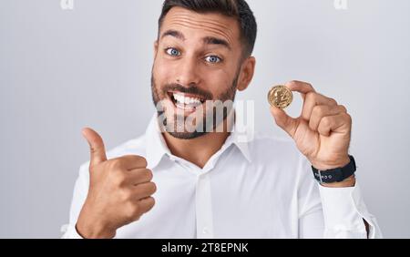 Bel homme hispanique tenant la pièce de monnaie de crypto-monnaie litecoin souriant heureux et positif, pouce vers le haut faisant excellent et signe d'approbation Banque D'Images