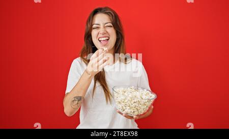 Jeune belle femme hispanique regardant un film de comédie mangeant du pop-corn riant beaucoup sur fond rouge isolé Banque D'Images