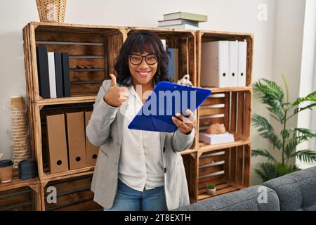Femme hispanique travaillant au bureau de consultation souriant heureux et positif, pouce vers le haut faisant excellent et signe d'approbation Banque D'Images