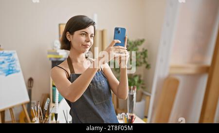 Détendue mais concentrée jeune, belle artiste hispanique, avec les cheveux courts, debout dans son atelier d'art, prenant une photo parfaite avec son smartphone Banque D'Images