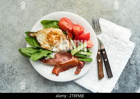 D'en haut de l'arrangement de petit déjeuner équilibré sur une assiette blanche avec oeuf au plat, bacon croustillant, feuilles d'épinards frais, concombre tranché et tomates en coin Banque D'Images