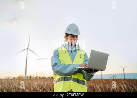 Femme ingénieure souriante en gilet de sécurité et en casque dur utilisant un ordinateur portable tout en se tenant dans un champ de parc éolien au coucher du soleil. Banque D'Images