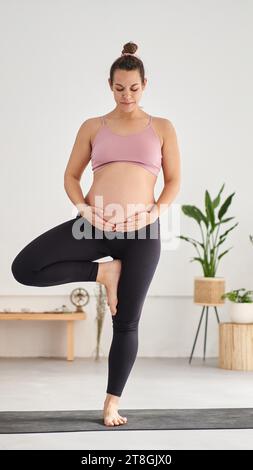 Portrait d'une femme caucasienne enceinte méconnaissable en vêtements de sport faisant une pose de yoga à l'intérieur. Banque D'Images