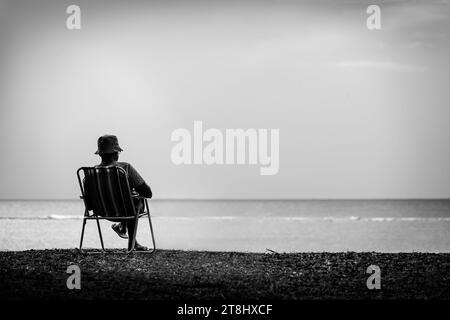 Image artistique en noir et blanc d'un homme solitaire assis sur une chaise de camping près de la plage et lisant un livre. Banque D'Images