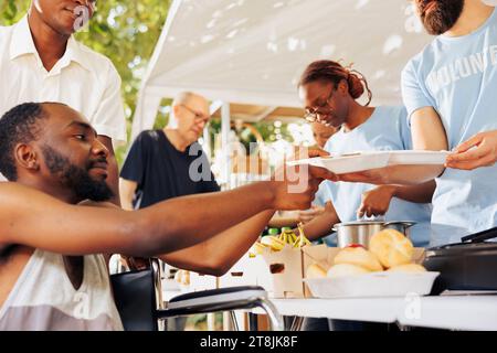 Le gars serviable aide et fournit de la nourriture gratuite à un homme pauvre afro-américain dans le besoin en fauteuil roulant. Les bénévoles d'une banque alimentaire extérieure fournissent une aide à la faim aux personnes infirmes et défavorisées. Banque D'Images