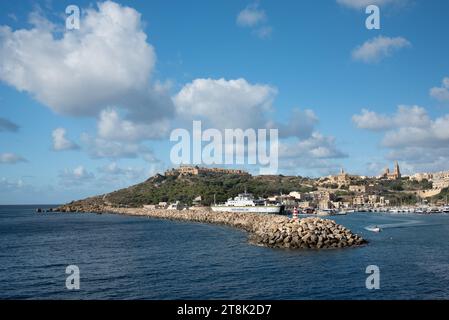 Vue sur le port de Gozo depuis le bord de mer. Un ferry est ancré dans le port. En arrière-plan se trouve la ville de Mgarr avec ses maisons et ses églises. Banque D'Images