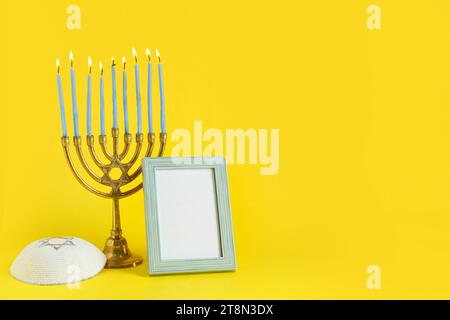 Menorah avec des bougies allumées, cadre d'image vierge et kippah pour la célébration Hanukkah sur fond jaune Banque D'Images