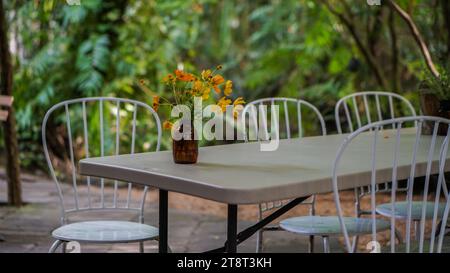 Chaise blanche moderne et table installée dans le jardin. Table à manger dans le jardin extérieur décoré avec des coquelicots de fleurs jaunes dans un vase en verre. Flou arrière Banque D'Images