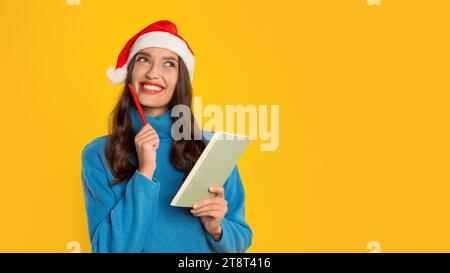Femme rêvant d'écrire des rêves festifs de Noël sur fond de studio jaune Banque D'Images