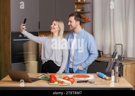 Un couple souriant et aimant prépare une salade ensemble tout en se tenant dans une cuisine à la maison et en utilisant un téléphone portable. Couple mignon homme et femme prenant la photo de foo Banque D'Images