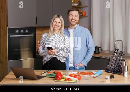 Un couple souriant et aimant prépare une salade ensemble tout en se tenant dans une cuisine à la maison et en utilisant un téléphone portable. Couple mignon homme et femme prenant la photo de foo Banque D'Images