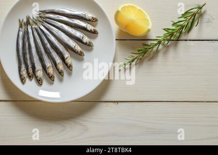 Anchois européen, hamsa frais, plusieurs petits poissons disposés sur assiette, romarin, citron, sur fond de planche de bois blanc, vue de dessus, espace pour copier le texte. Banque D'Images