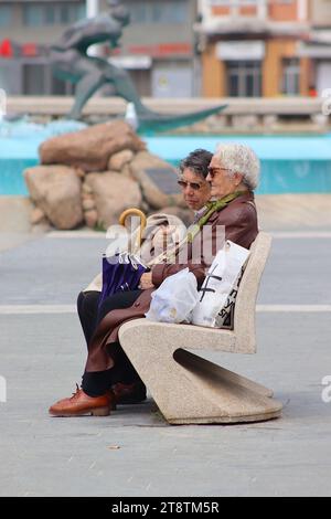 Deux dames âgées reposant en conversation sur un banc public à côté de la fontaine de Los Surfistas sur le hall de la plage de Orzán, la Corogne, Espagne. Banque D'Images