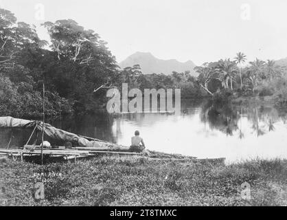 Canoë fidjien sur les rives de la rivière Lami, Viti Levu, Fidji, vue d'un canoë fidjien avec voile à fourrure au bord de l'eau sur la rivière Lami. Un homme est assis sur le canot et regarde la rivière. Les palmiers poussent sur la rive opposée et il y a des montagnes au loin. Prise au début des années 1900 Banque D'Images