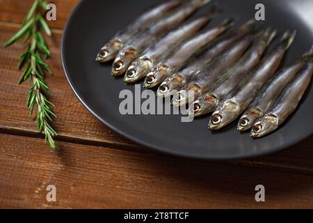 Anchois européen, hamsa frais, plusieurs petits poissons disposés en rangée sur plaque grise, romarin, sur fond de table en planche marron en bois, vue de côté, selec Banque D'Images