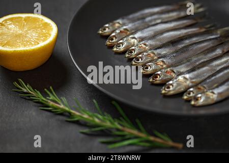 Anchois européen, hamsa frais, plusieurs petits poissons romarin et citron sur plaque grise sur fond de table en planche marron en bois, vue de dessus, espace à copier Banque D'Images