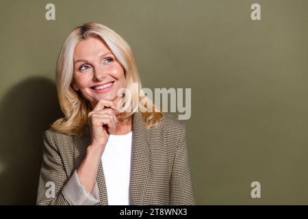 Photo de dame souriante mature dans la veste toucher menton regardant la nouveauté hmm pensées imaginer ses plans isolés sur fond de couleur kaki Banque D'Images
