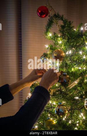 Mains féminines attachant des lumières et des boules à l'arbre de Noël à l'intérieur. Préparations de Noël et décoration d'arbre pour la saison des fêtes Banque D'Images