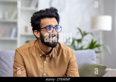 Photo en gros plan d'un jeune Indien dans des lunettes assis sur le canapé à la maison et regardant sur le côté avec un sourire. Banque D'Images