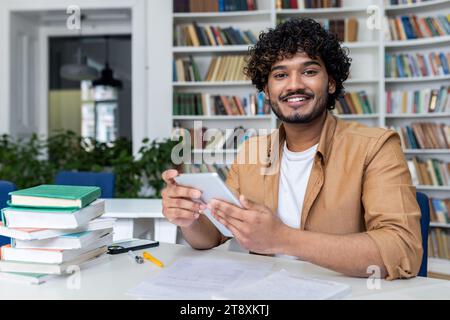 Portrait d'un jeune étudiant indien assis dans la bibliothèque de l'université, tenant une tablette et souriant à la caméra. Banque D'Images