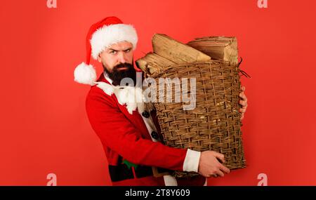 Homme barbu en costume de Père noël portant un panier de bois de chauffage haché pour cheminée. Vacances d'hiver. Père Noël avec bois de chauffage en osier Banque D'Images