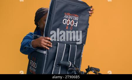 Femme de livraison opportune arrivant avec le quartier urbain de vélo et déchargeant la nourriture rapide à emporter. Chez le client, le courrier afro-américain féminin ouvre son sac de livraison de nourriture. Banque D'Images