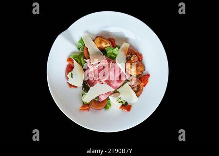 Salade italienne avec jambon cru, tomate et parmesan dans un plat blanc rond sur fond noir, vu de dessus. Banque D'Images