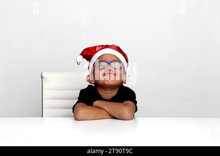 Un garçon latino de 7 ans à la peau foncée avec un chapeau de Noël du Père Noël a l'air triste et en colère parce qu'il n'aime pas les vacances et qu'ils ne lui ont pas donné Banque D'Images