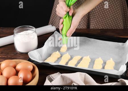 Faire des eclairs. Femme pressant la pâte crue du sac à pâtisserie sur le plateau de cuisson à la table en bois, closeup Banque D'Images