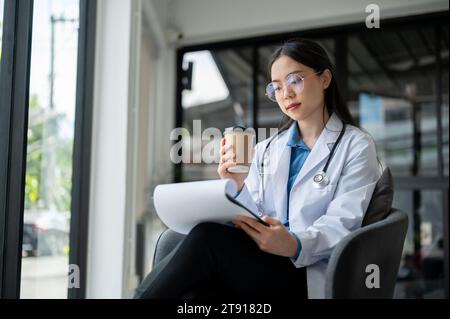 Une médecin asiatique professionnelle et expérimentée lit et examine ses cas médicaux sur un presse-papiers médical dans son bureau. Conceps de santé Banque D'Images