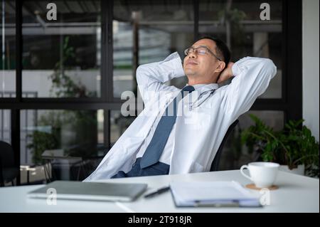 Un médecin asiatique senior détendu et insouciant est appuyé sur sa chaise, mettant ses mains derrière sa tête et faisant une sieste, reposant dans son bureau. Banque D'Images