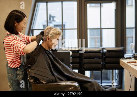 Portrait de vue latérale de jeune homme asiatique souriant dans le salon de beauté avec coiffeur appliquant de l'eau de Javel sur les cheveux, espace de copie Banque D'Images