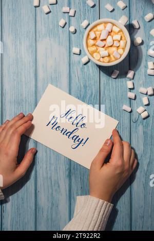 BONJOUR LUNDI bleu lundi texte concept main féminine tenant la carte avec tasse blanche de café et guimauves sur fond en bois. Le jour le plus déprimant de l'année en janvier. Jour commettre suicide et signe de motivation de dépression. Vue de dessus, pose à plat Banque D'Images