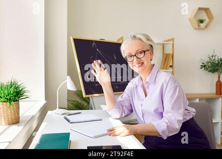 Portrait de la femme senior amicale professeur de mathématiques en ligne assis dans la salle de classe devant la caméra Web. Banque D'Images