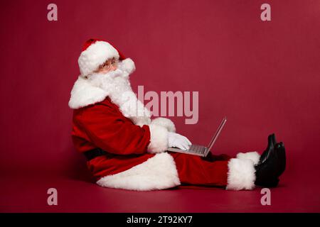 Vue latérale photo de joyeux Père Noël souriant utilise un ordinateur portable sur un fond rouge Banque D'Images